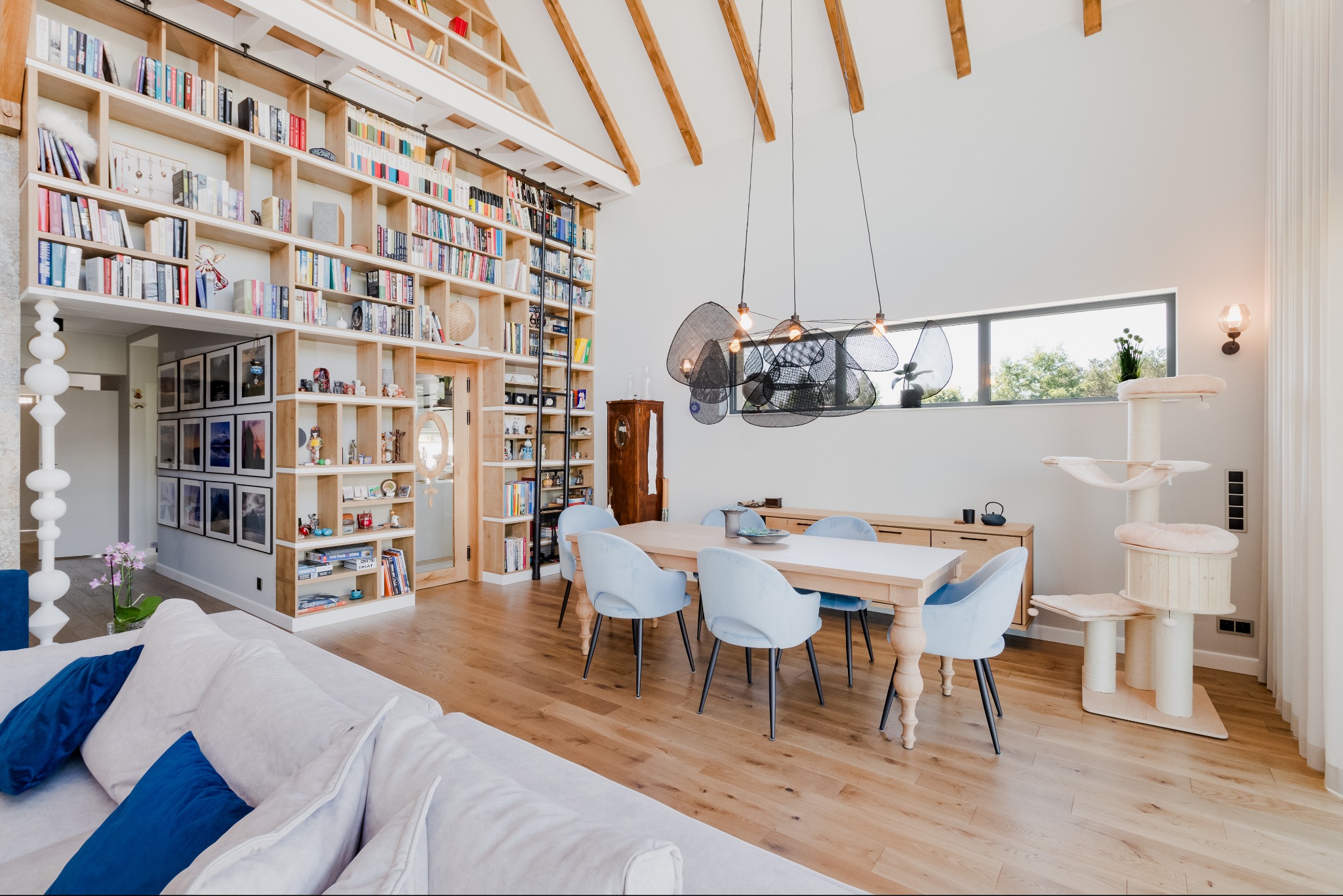 Jasne wnętrze domu z wysokim sufitem i drewnianymi belkami, duży regał z książkami, stół z jasnymi krzesłami, designerska lampa wisząca i drapak dla kota
