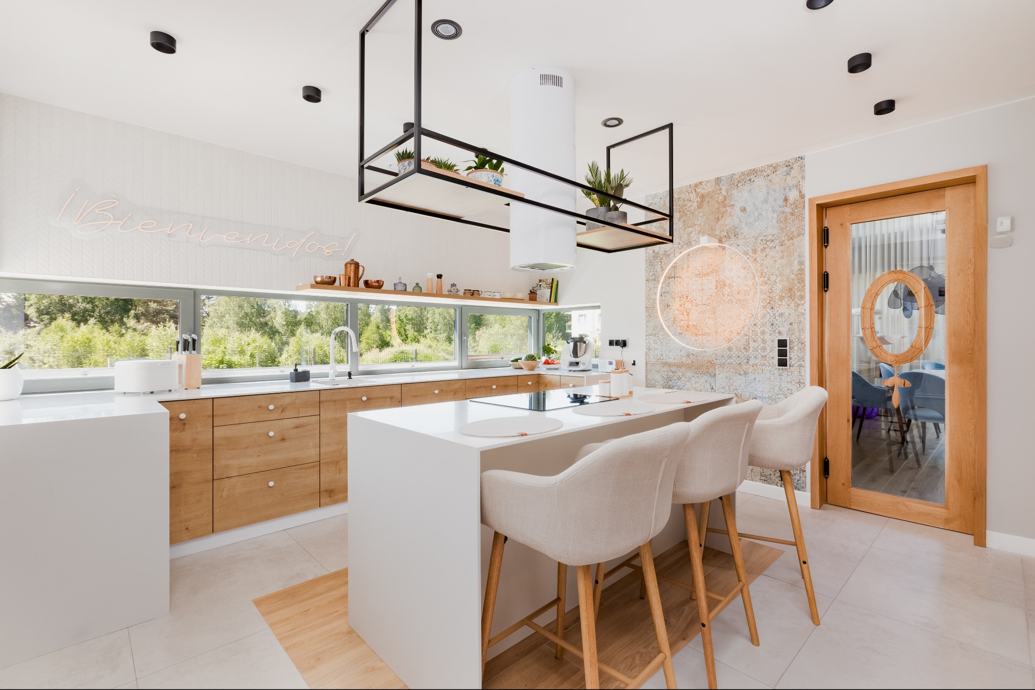 Jasna, nowoczesna kuchnia z wyspą, drewnianymi szafkami i dużym oknem z widokiem na zieleń, oświetlona neonowym napisem 'Bienvenidos!'