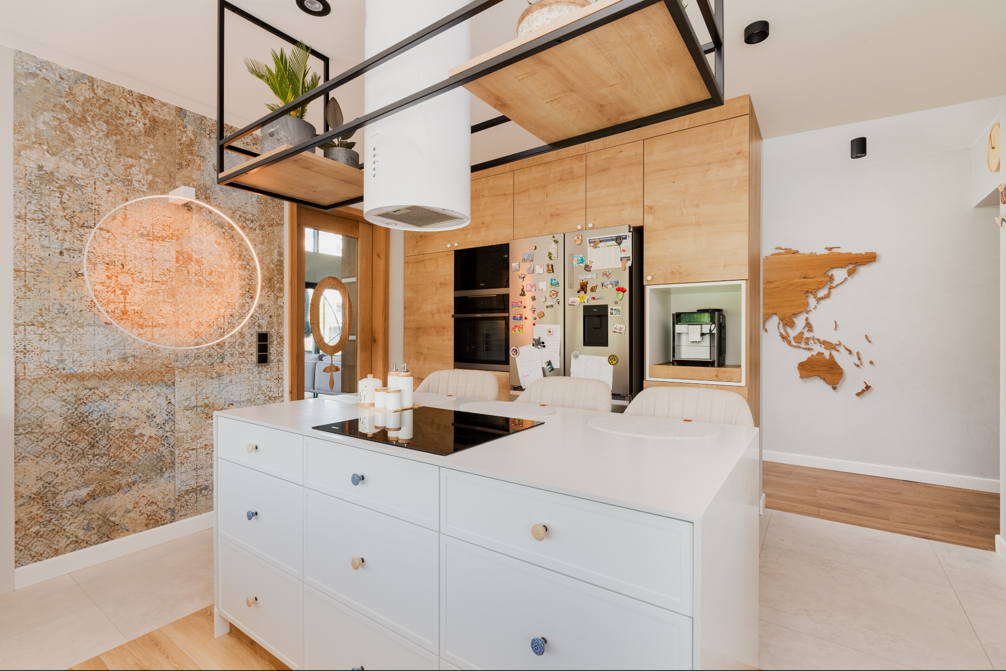 Jasna kuchnia w Gdańsku z białą wyspą, drewnianymi szafkami, i dekoracyjną mapą świata na ścianie, oświetlona okrągłą lampą.