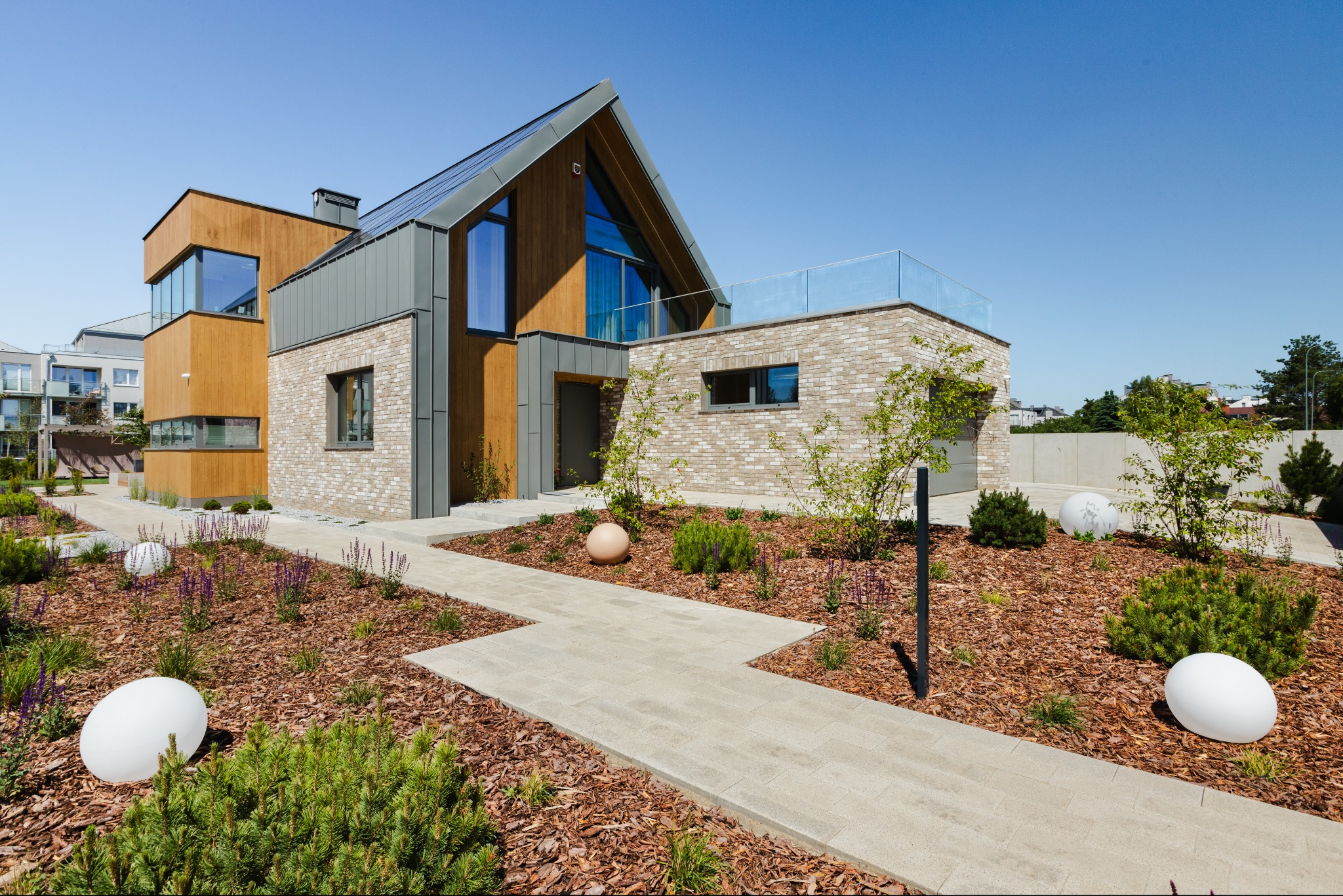 Nowoczesny dom jednorodzinny z drewnianą elewacją, ceglaną ścianą i szarymi elementami wykończeniowymi, otoczony ogrodem z ścieżką z kostki i kulistymi lampami.