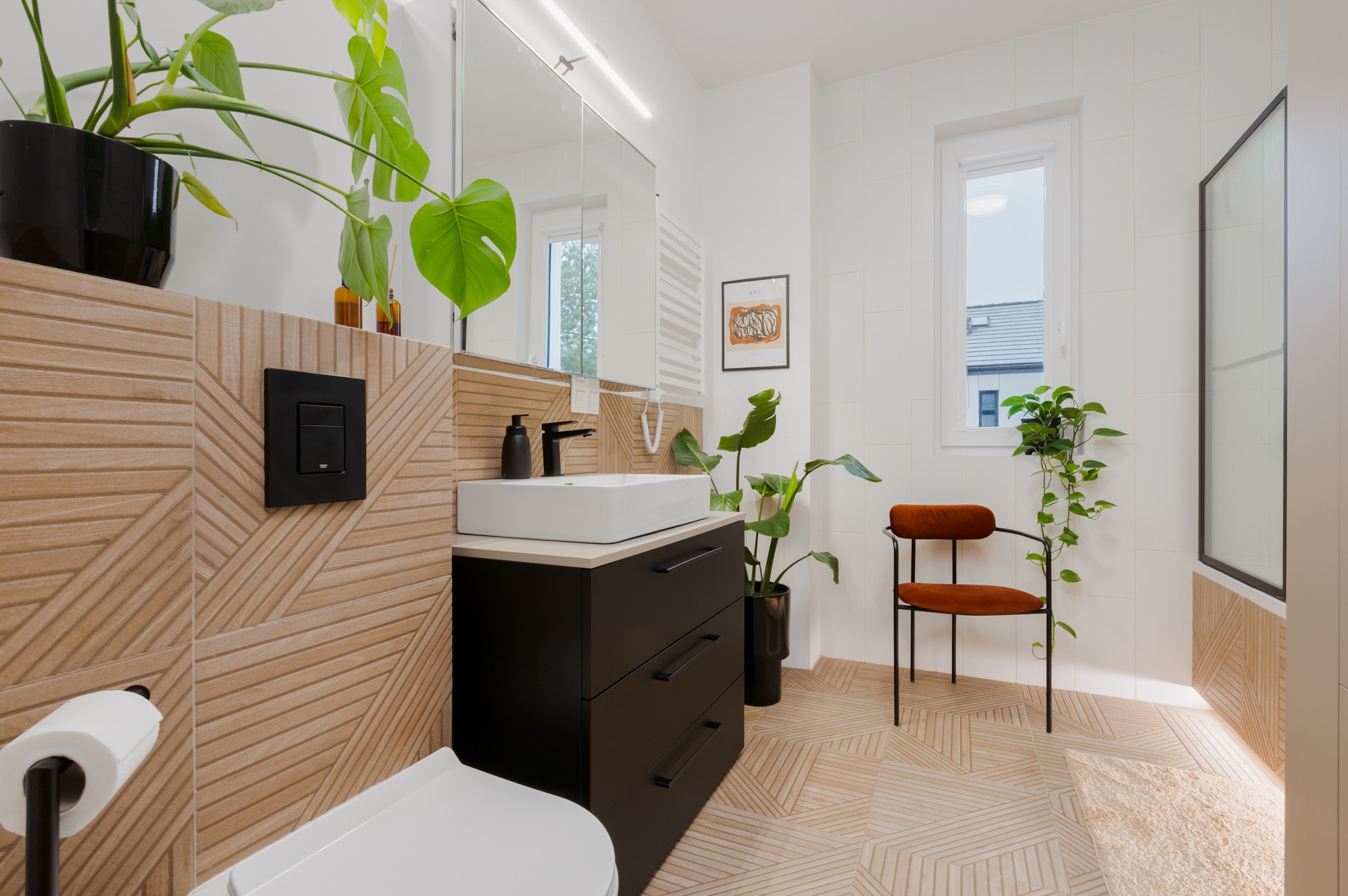 Nowoczesna łazienka z geometrycznymi płytkami na ścianach i podłodze, białą umywalką na czarnej szafce, roślinami doniczkowymi i krzesłem z pomarańczowym siedziskiem.