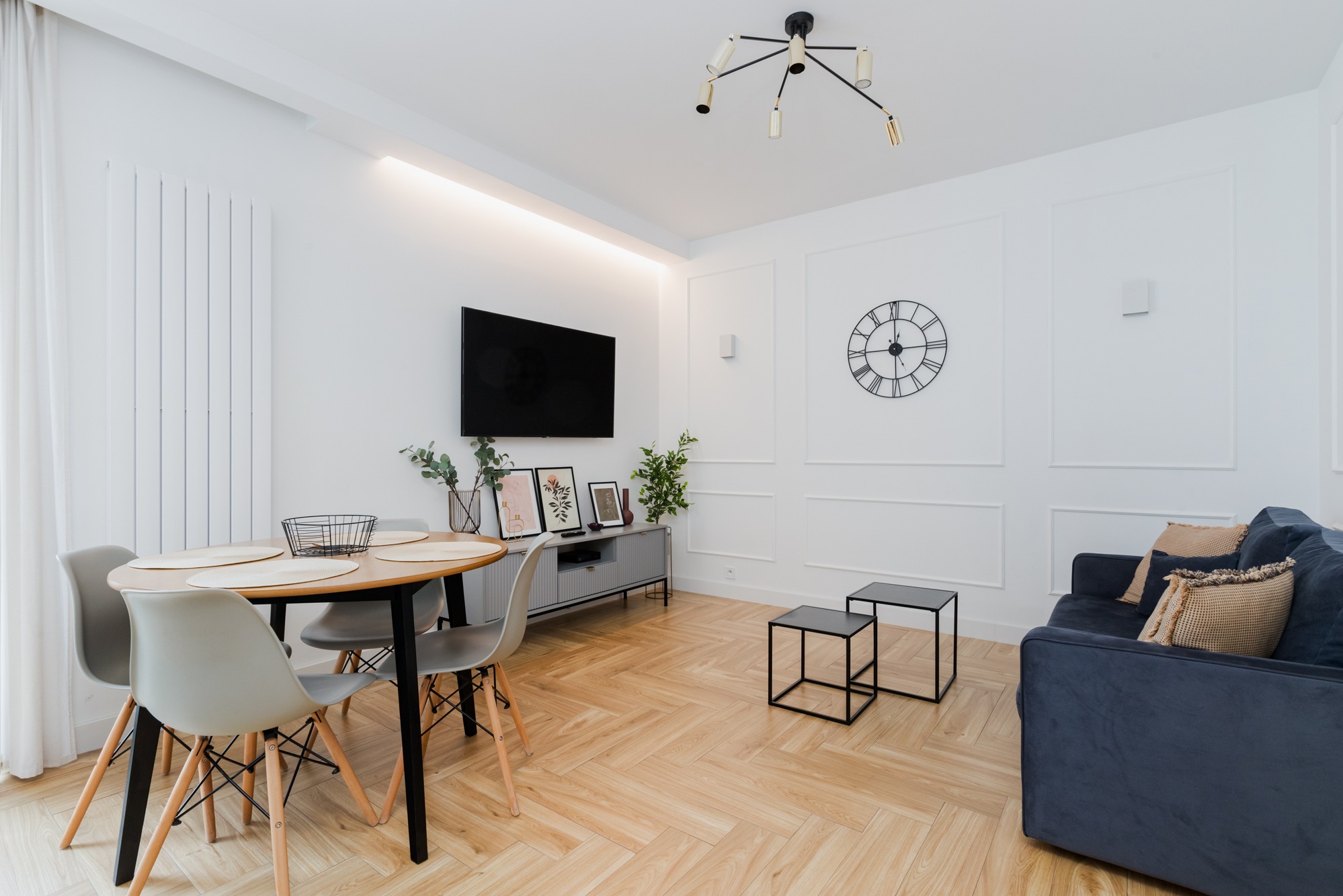 Jasne wnętrze salonu z drewnianą podłogą w jodełkę, okrągłym stołem z szarymi krzesłami, szarą komodą pod telewizorem, granatową kanapą i czarnym zegarem na ścianie.