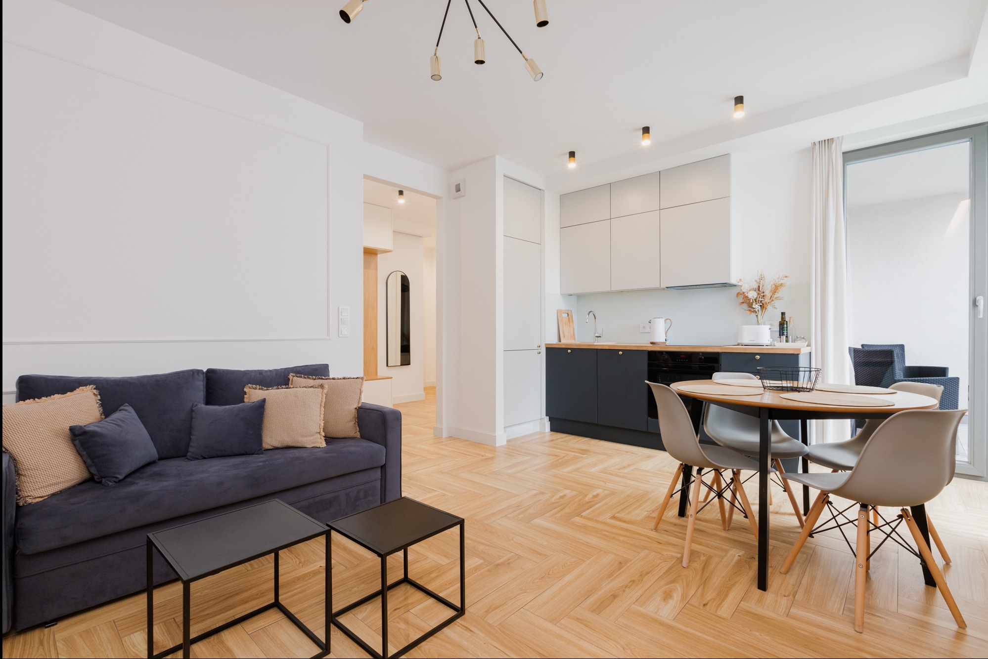 Jasne, przestronne wnętrze mieszkania z parkietem w jodełkę, szarą sofą z poduszkami, nowoczesną kuchnią z ciemnymi szafkami dolnymi i białymi górnymi oraz okrągłym stołem z krzesłami.