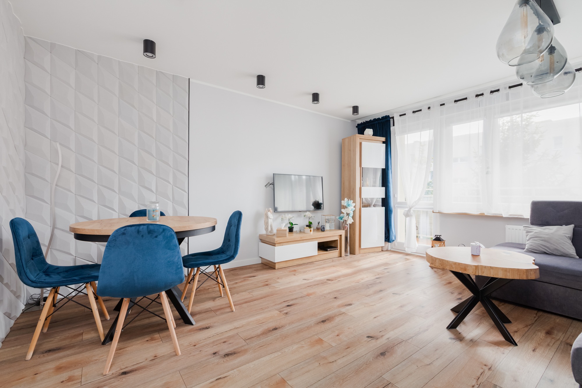 Jasne wnętrze salonu z drewnianą podłogą, okrągłym stołem z niebieskimi krzesłami, telewizorem na szafce RTV i stolikiem kawowym z blatem z naturalnego drewna.