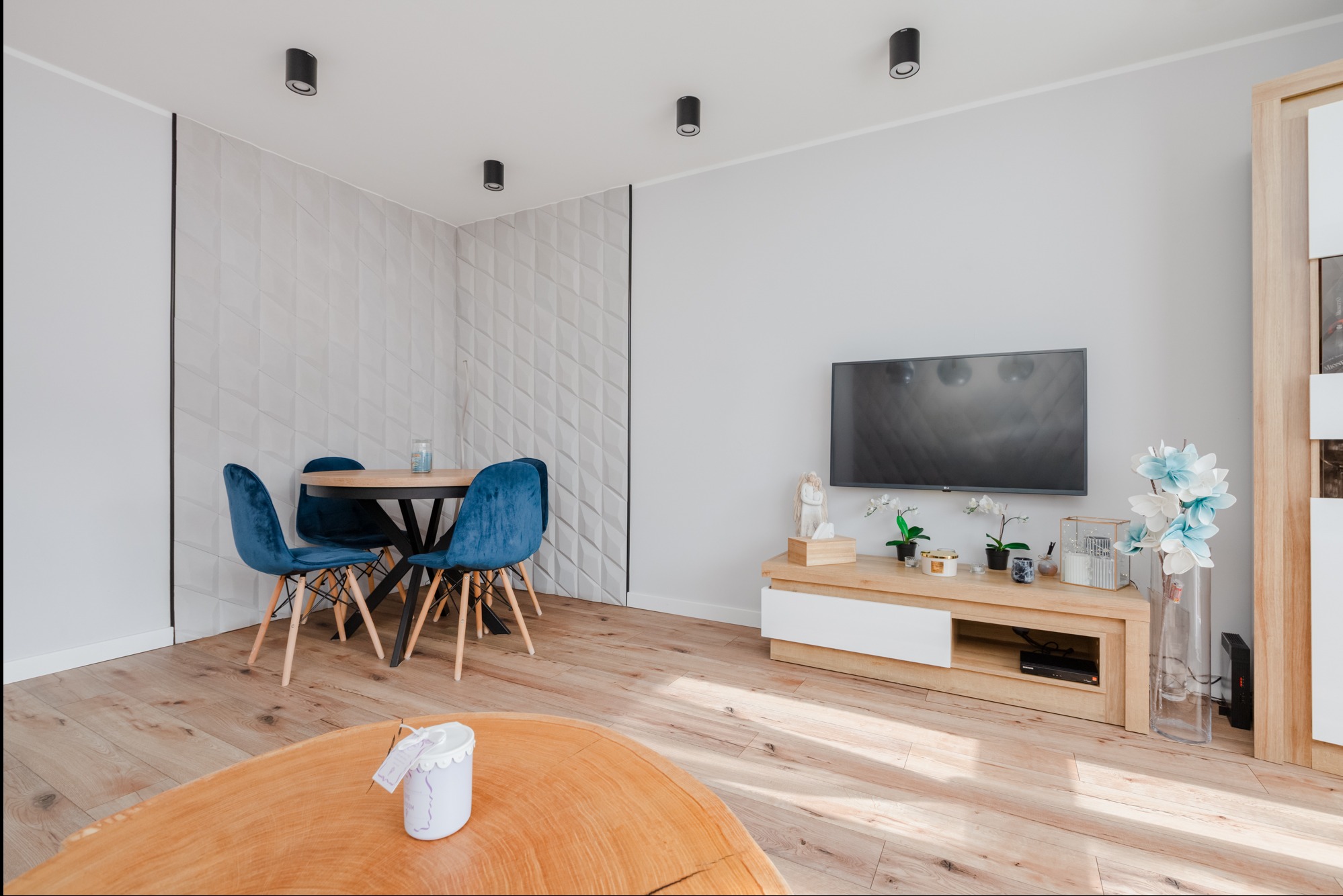 Jasne wnętrze salonu z drewnianą podłogą, okrągłym stołem z niebieskimi krzesłami, telewizorem na szafce RTV i dekoracyjną ścianą z trójwymiarowych paneli.