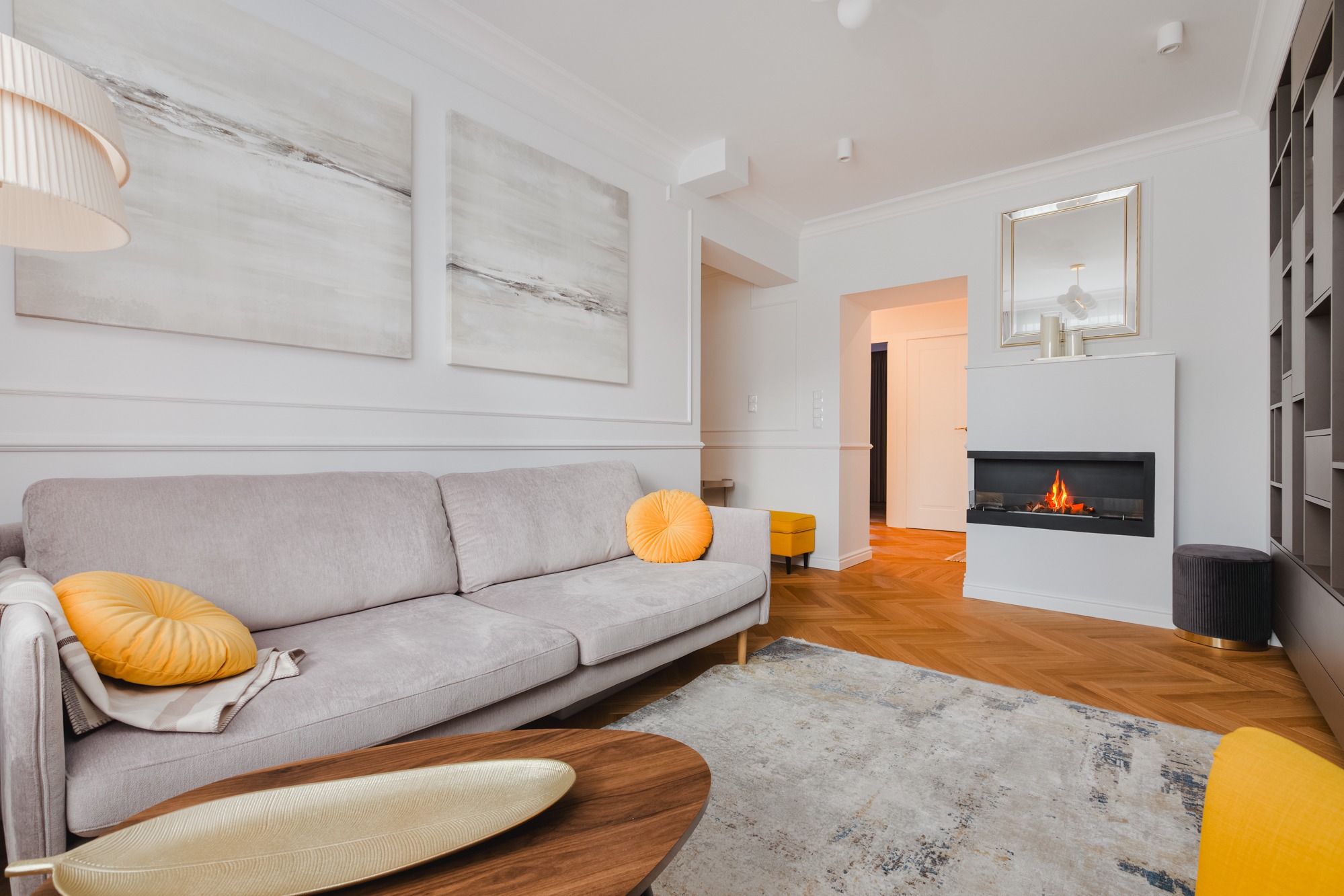 Elegancki salon z beżową sofą, okrągłymi żółtymi poduszkami, drewnianym stolikiem kawowym i kominkiem. Na ścianach abstrakcyjne obrazy i lustro w złotej ramie.