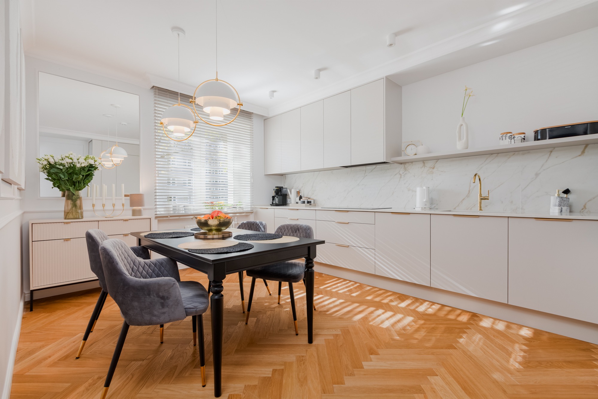 Jasna kuchnia z jadalnią: czarny stół z szarymi krzesłami, parkiet w jodełkę, białe szafki z marmurowym blatem i złotą armaturą. Nowoczesne lampy wiszące.
