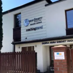 Dwupiętrowy budynek z białymi ścianami i brązowymi ramami okiennymi, z szyldami firmowymi 'Norman Benett Group' i 'Coaching Travel' na górze oraz 'Dobra Aura Gabinet Masażu' na dole, widziany zza...