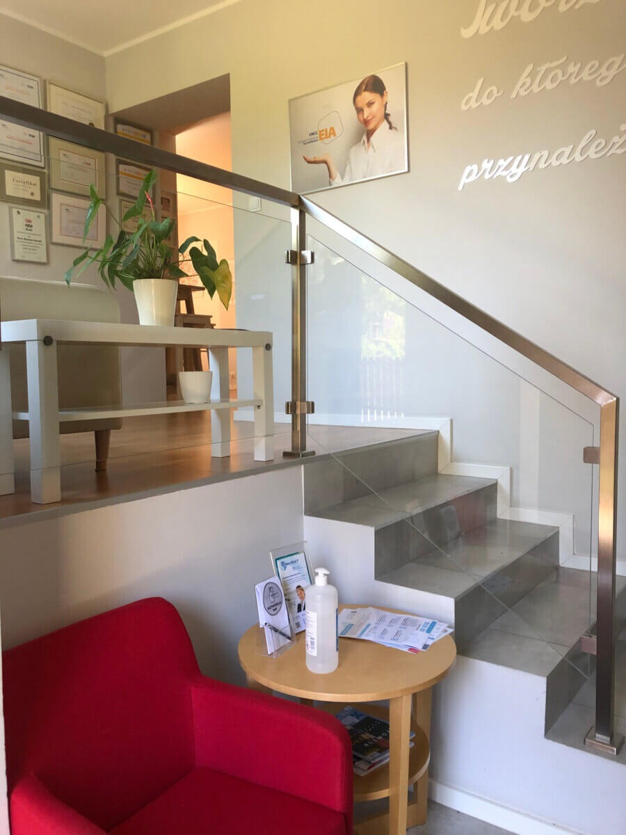 Jasne wnętrze poczekalni kliniki: czerwony fotel, stolik z płynem do dezynfekcji, schody z szklaną balustradą, dyplomy na ścianie i plakat z wizerunkiem kobiety.