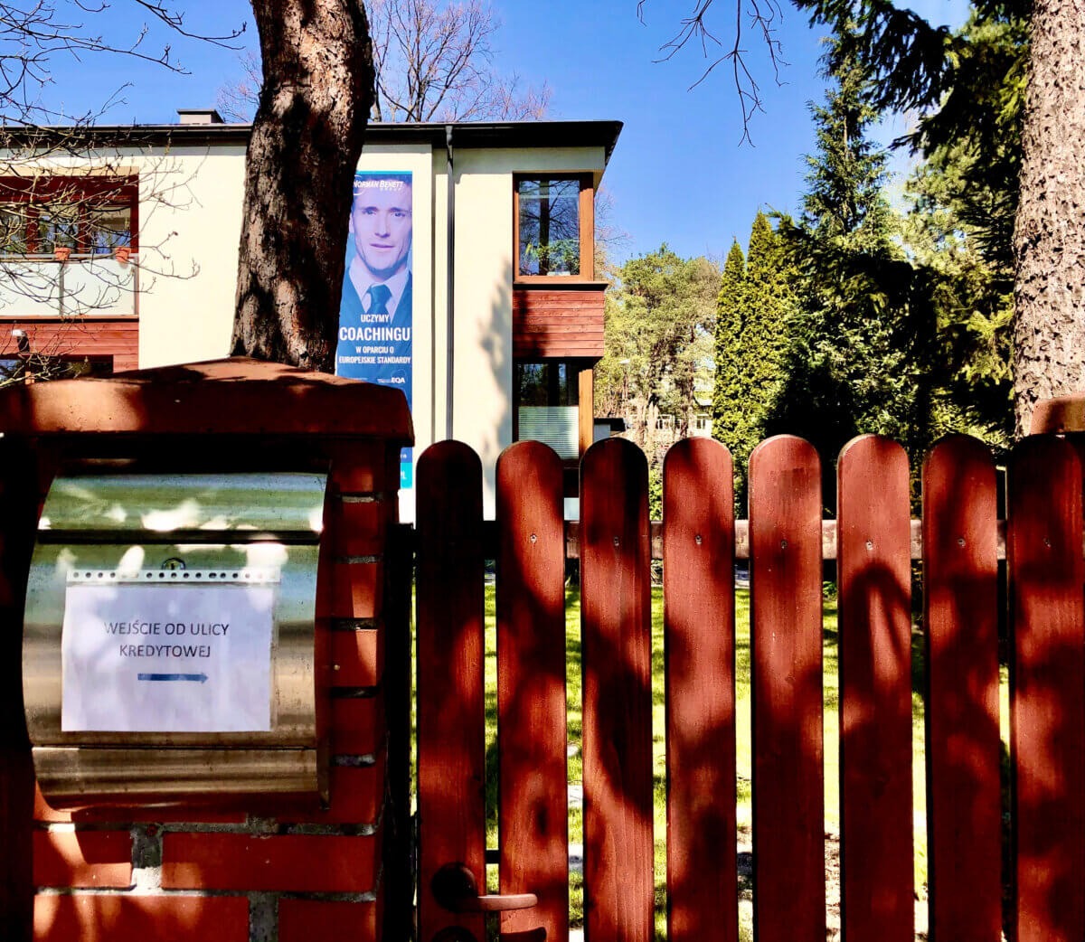 Budynek z banerem reklamującym coaching, widoczny zza drewnianego płotu i skrzynki pocztowej z kartką informacyjną.