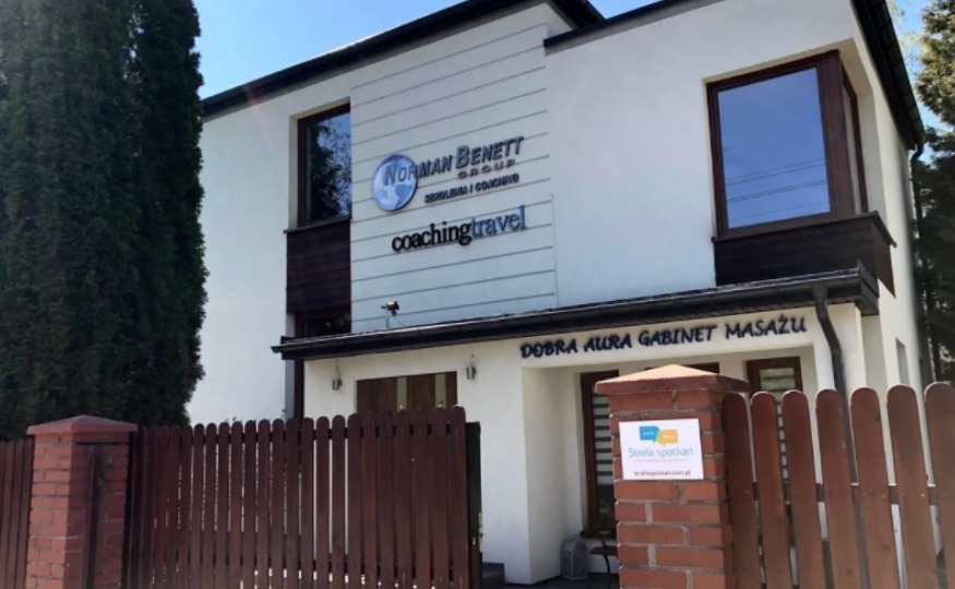 Dwupiętrowy budynek z białymi ścianami i brązowymi ramami okiennymi, z szyldami firmowymi 'Norman Benett Group' i 'Coaching Travel' na górze oraz 'Dobra Aura Gabinet Masażu' na dole, widziany zza...