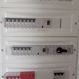 Rozdzielnica elektryczna z wieloma bezpiecznikami, w tym zabezpieczeniami przeciwprzepięciowymi oznaczonymi numerami od 1 do 57, zamontowana na białej ścianie.