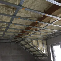 Poddasze z ociepleniem pianą PUR, widoczne metalowe profile konstrukcji sufitu podwieszanego, fragment drewnianych belek stropowych i przewód elektryczny.