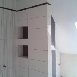 Wykończona ściana w łazience z białych płytek z ciemnymi fugami i wnękami. Ciemny pas płytek na g&oacute;rze. Częściowo widoczna ściana z pionowymi paskami.