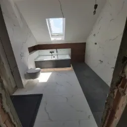 Nowoczesna łazienka na poddaszu z oknem dachowym, wykończona białymi i szarymi płytkami imitującymi marmur, z wiszącą toaletą i wanną obudowaną ciemnoszarą zabudową, widok z dołu w trakcie prac...