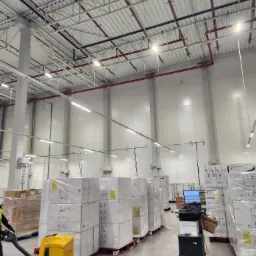 Oświetlenie LED w magazynie wysokiego składowania z widocznymi instalacjami pod sufitem, rurami przeciwpożarowymi i lampami liniowymi.