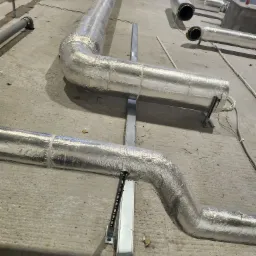 Izolowane rury wentylacyjne zamontowane na metalowych wspornikach w surowym wnętrzu, widoczne kable i resztki materiałów budowlanych na podłodze.