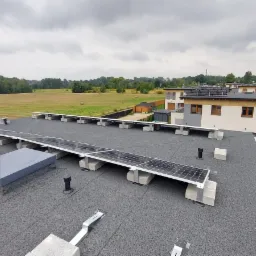 Widok z góry na dach pokryty papą z zainstalowanymi panelami fotowoltaicznymi na betonowych podstawach oraz klimatyzator, w tle budynki mieszkalne i pole z belami siana.