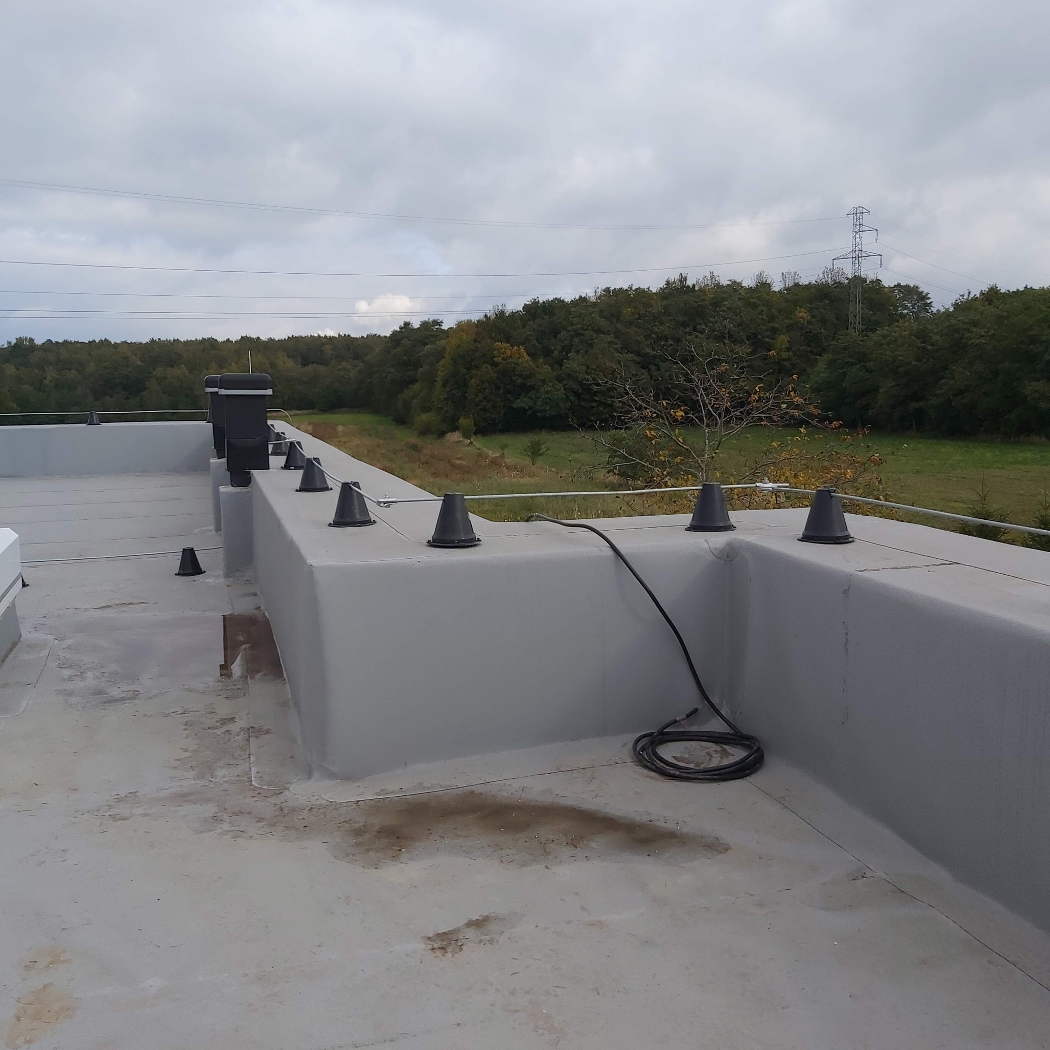 Płaski dach budynku z zamontowanymi odgromnikami stożkowymi i przewodami uziemiającymi, w tle las i słup wysokiego napięcia, pochmurne niebo.