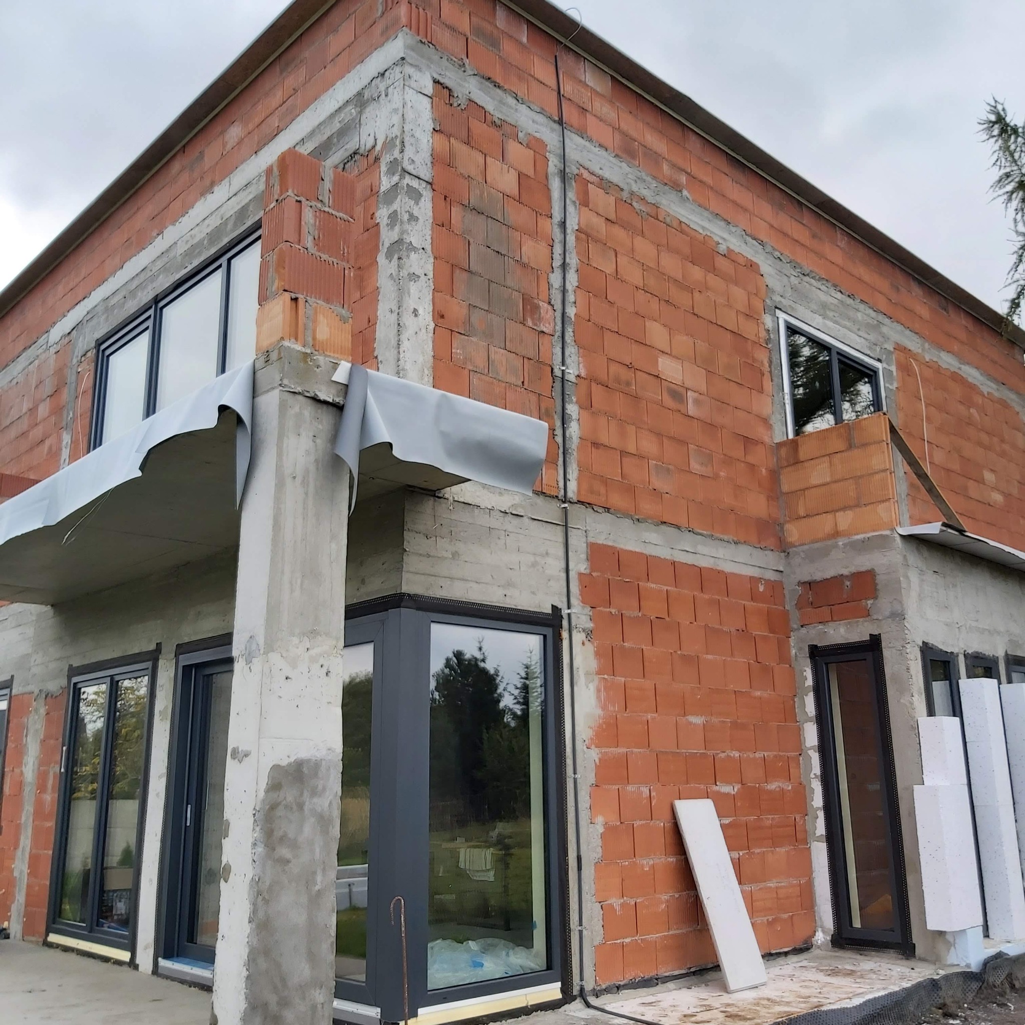 Surowy stan budynku w trakcie budowy: widoczne cegły, betonowe filary, okna z czarnymi ramami, wystające przewody elektryczne na zewnątrz budynku.