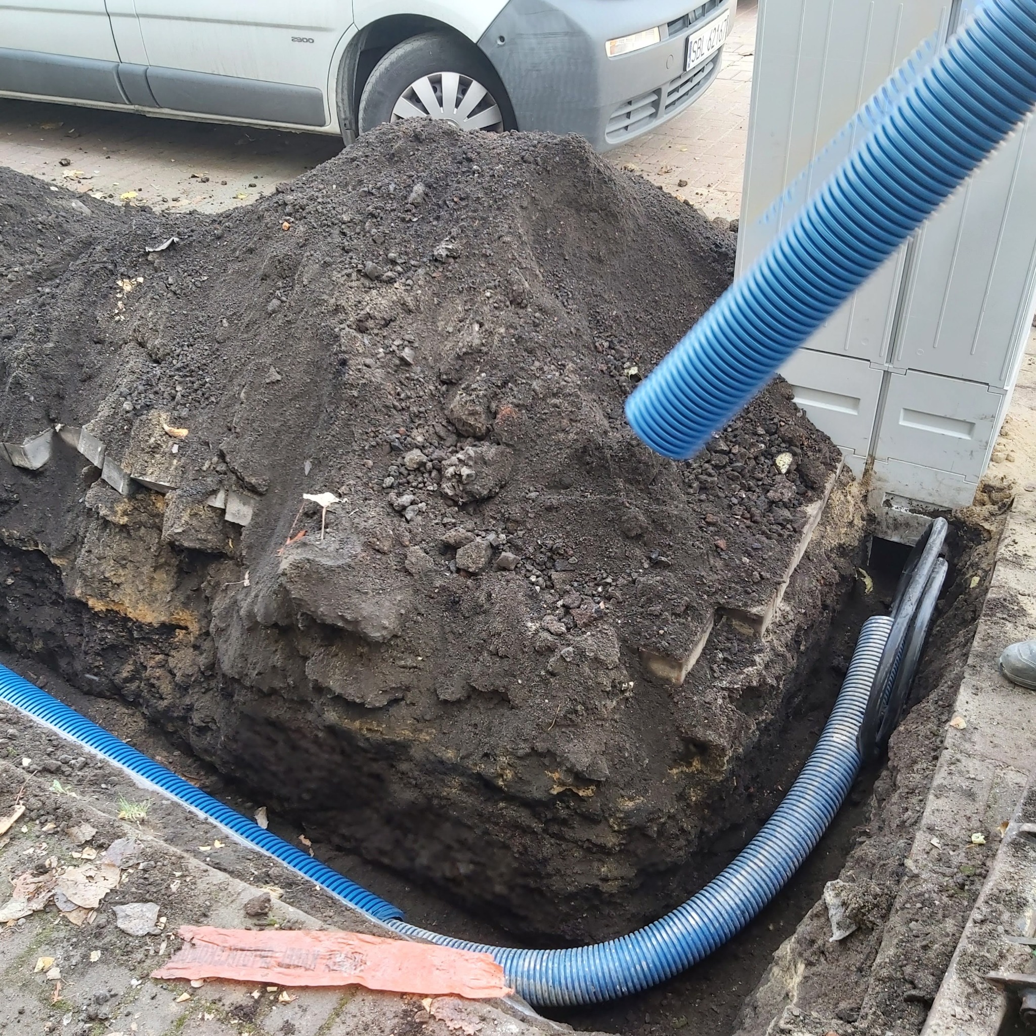 Wykop z ułożonymi niebieskimi rurami karbowanymi, prawdopodobnie peszel, przyłącze do skrzynki elektrycznej, widoczny fragment białego samochodu dostawczego.