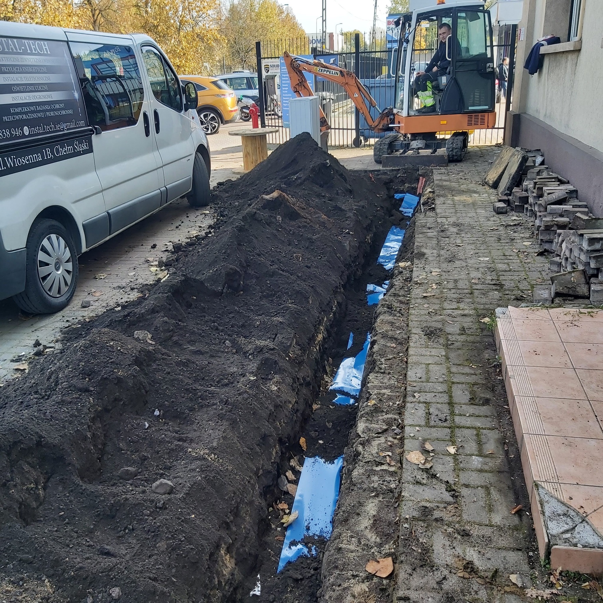 Wykop z niebieskimi rurami w ziemi, obok zaparkowany biały bus z napisem INSTAL-TECH oraz pomarańczowa mini koparka Zeppelin z operatorem w środku, sterta kostki brukowej i ziemi.