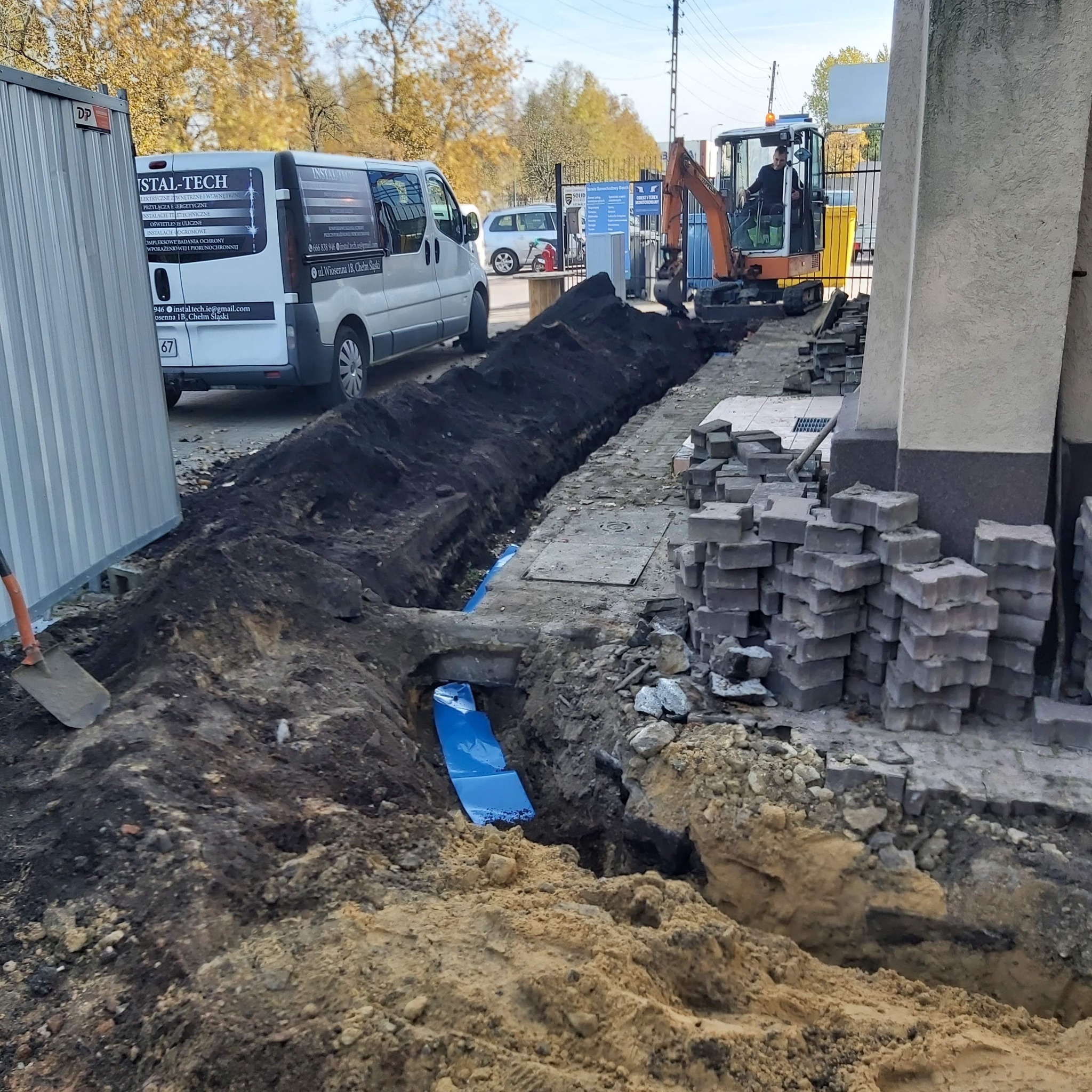 Wykop pod przyłącze, widoczna folia zabezpieczająca w wykopie, obok zaparkowany bus z napisem Instal-Tech, na drugim planie koparka.
