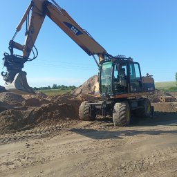 PROF-BRUK Radosław Mańkowski - Żółta koparka kołowa CAT M315C na placu budowy, z łyżką skierowaną w stronę pryzmy piasku, w tle zielone pole i błękitne niebo.