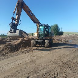 PROF-BRUK Radosław Mańkowski - Żółta koparka CAT na kołach nabiera piasek na placu budowy w słoneczny dzień, widoczne ślady opon i niewielkie kałuże.