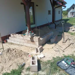 Budowa murku z betonowych bloczków przy domu z drewnianą konstrukcją dachu, widoczne narzędzia budowlane i poziomica.