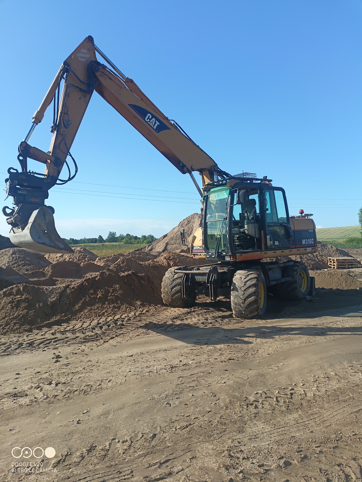Żółta koparka kołowa CAT M315C na placu budowy, z łyżką skierowaną w stronę pryzmy piasku, w tle zielone pole i błękitne niebo.