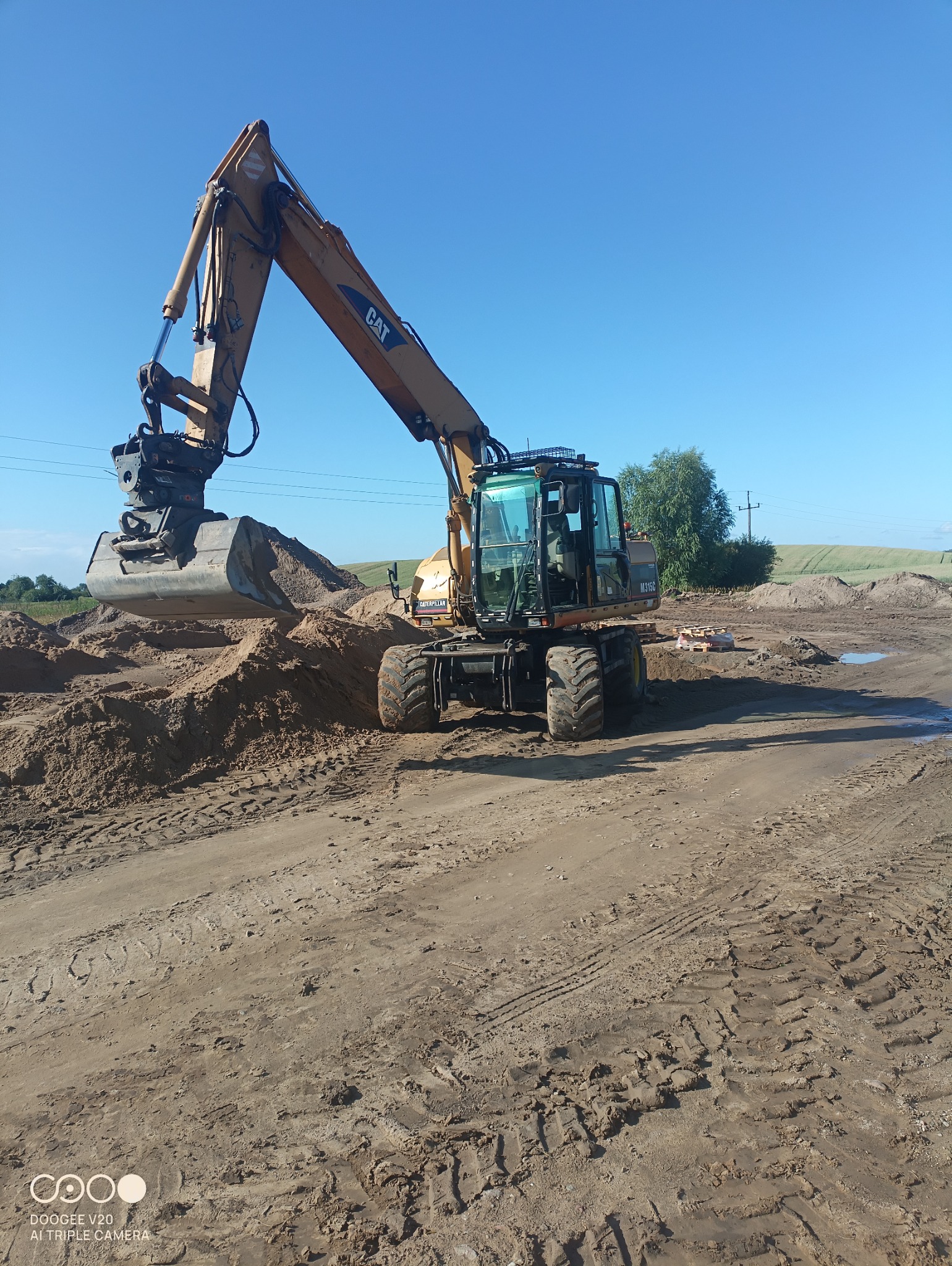 Żółta koparka CAT na kołach nabiera piasek na placu budowy w słoneczny dzień, widoczne ślady opon i niewielkie kałuże.