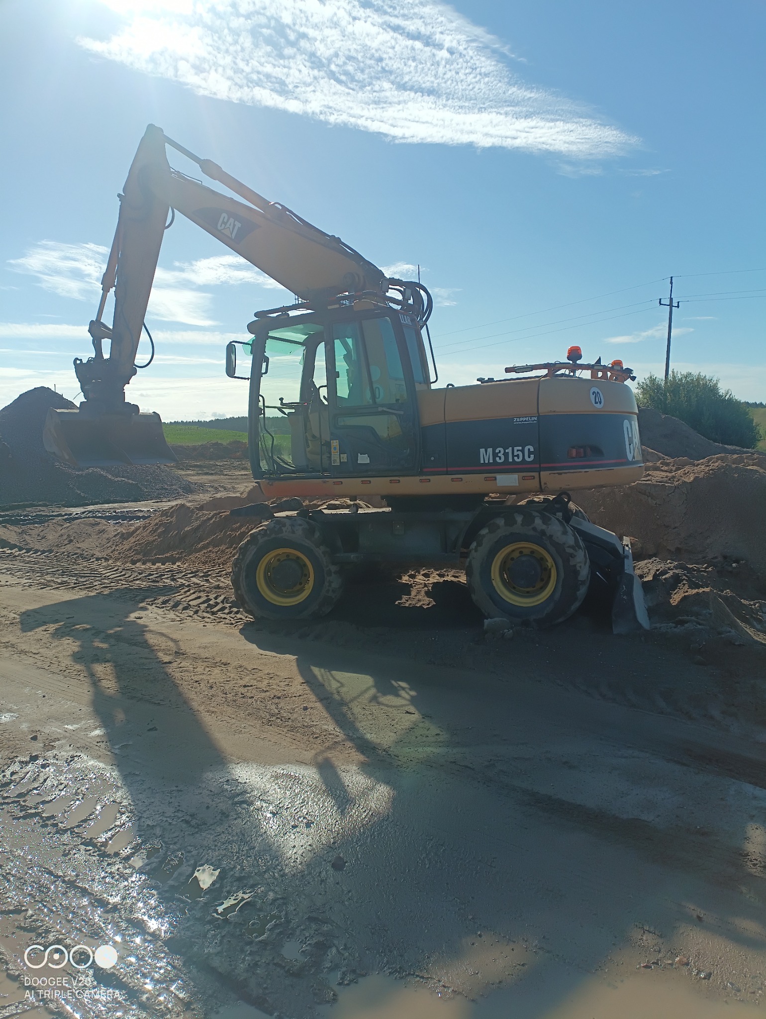 Żółta koparka kołowa CAT M315C nabiera piasek na placu budowy w słoneczny dzień, widoczne ślady opon w błocie.