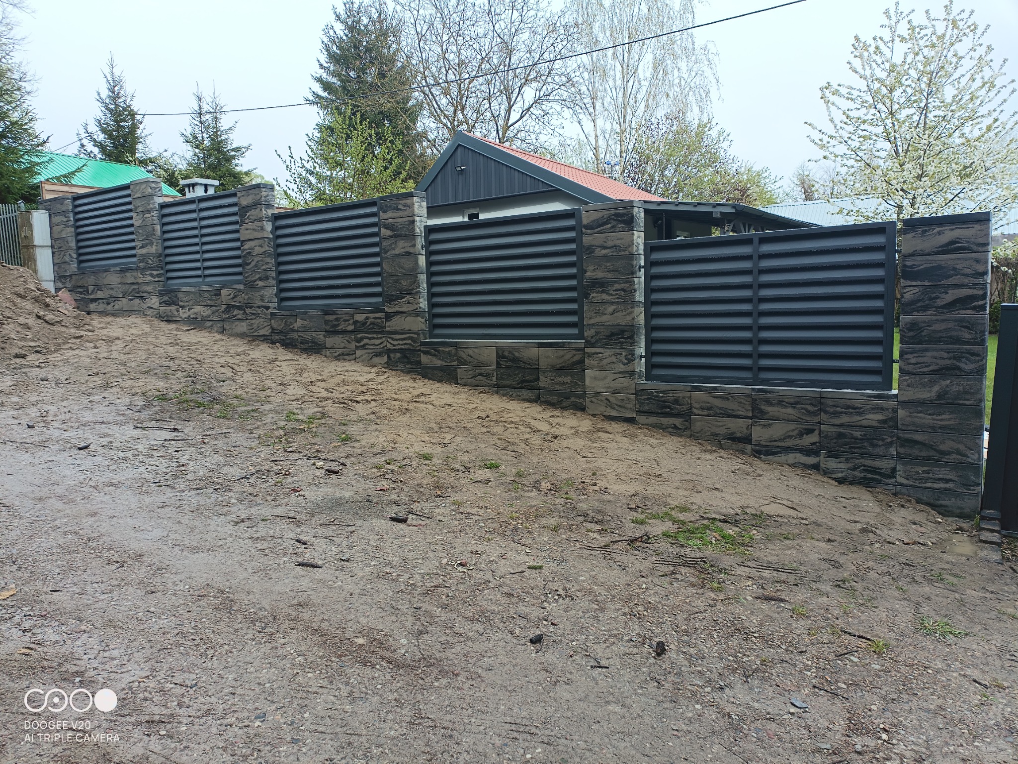 Nowoczesne, poziome ogrodzenie z szarych paneli metalowych i kamiennych słupków, widoczne z perspektywy terenu z ziemią i zielenią w tle.