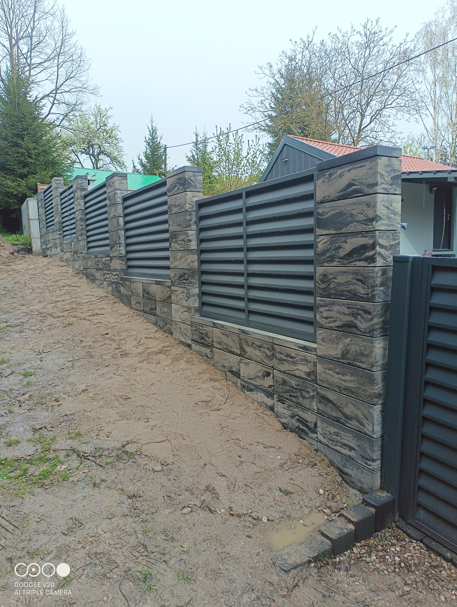 Nowoczesne ogrodzenie panelowe z betonowymi słupkami o marmurkowym wzorze, widoczne ślady błota po deszczu.