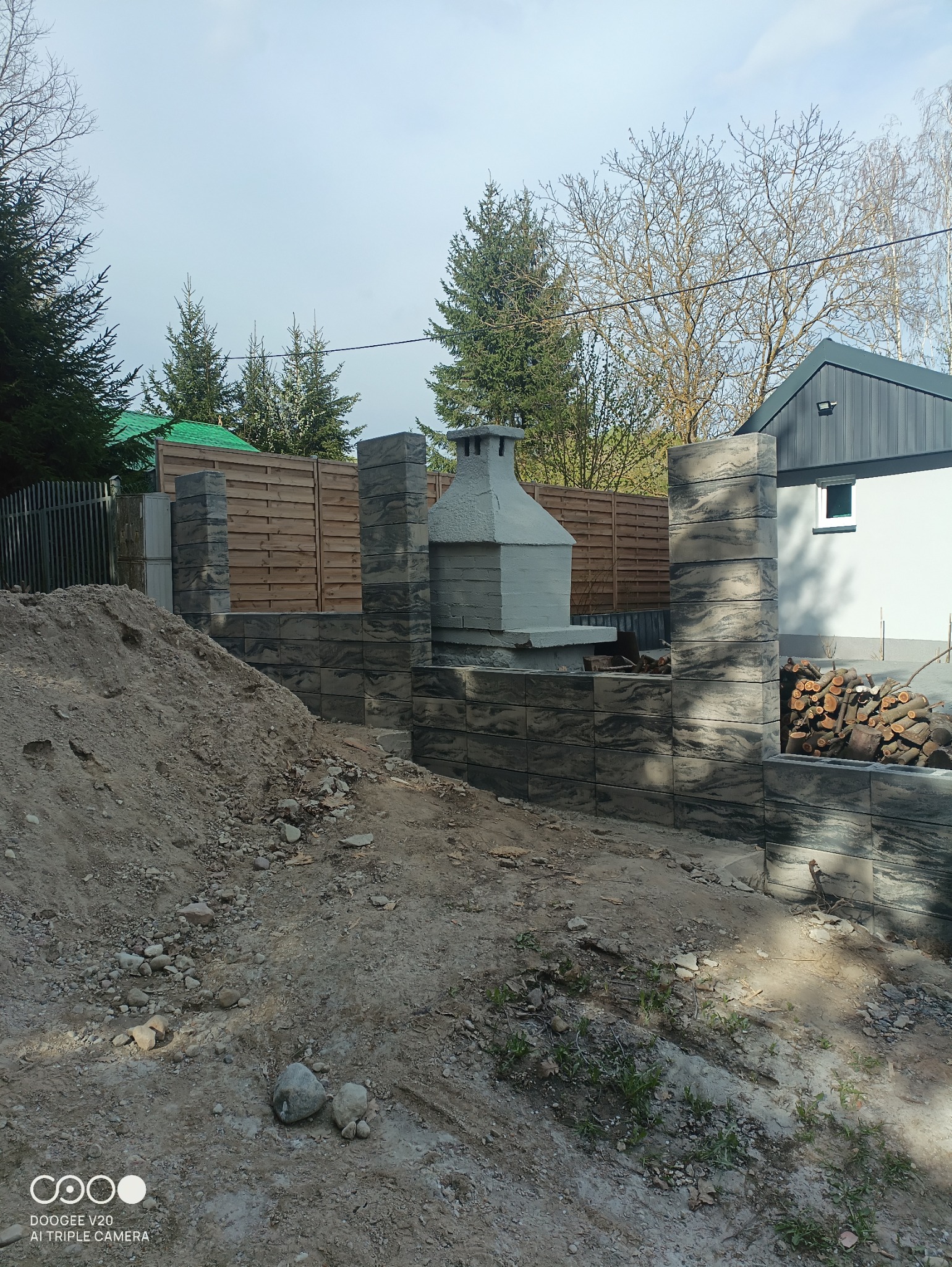 Budowa grilla ogrodowego z szarej kostki imitującej kamień, widoczny stos drewna, w tle drewniany płot i dom.