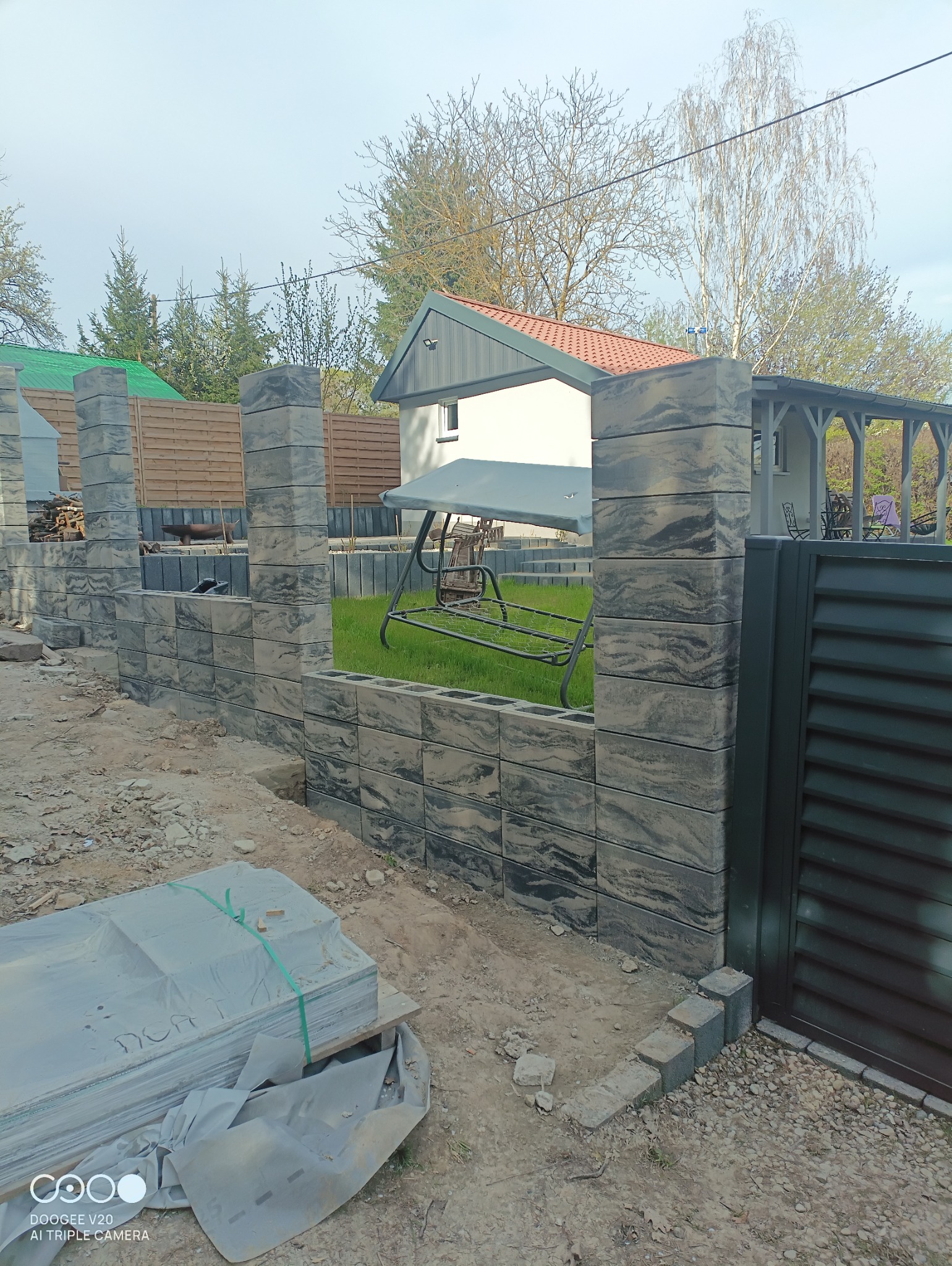 Wznoszenie ogrodzenia z betonowych bloczków o wzorze imitującym marmur, widoczne elementy furtki, w tle dom z ogrodem i huśtawką.