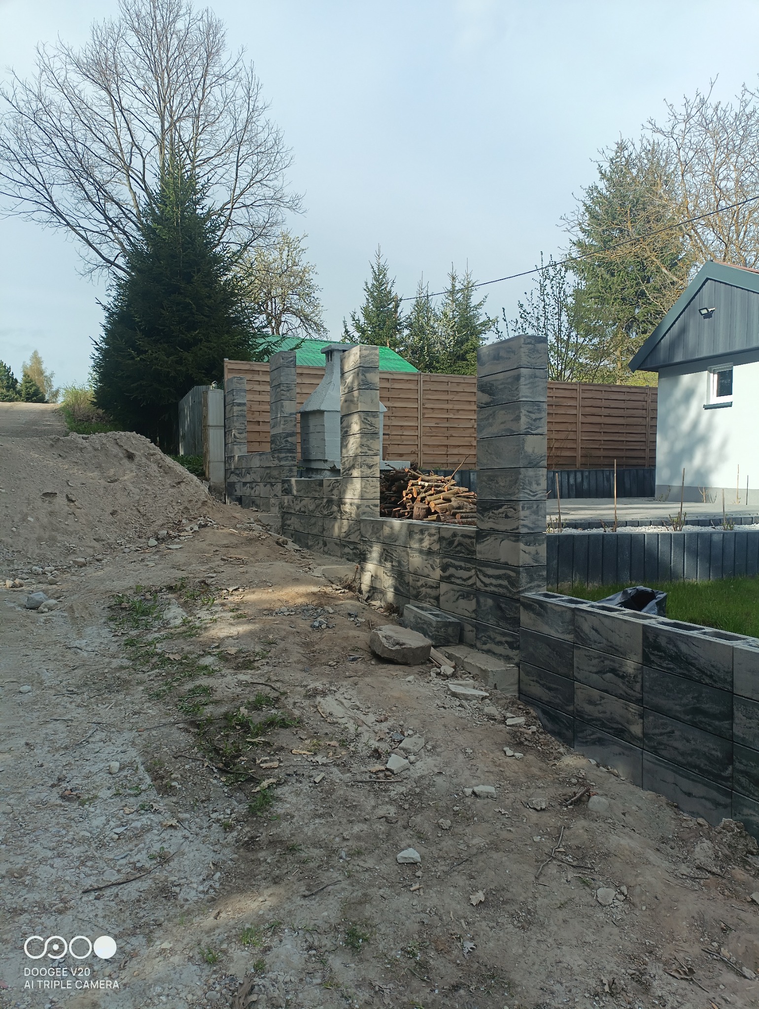 Wznoszona z szarych bloczków ogrodowa ściana z kominkiem i ułożonym drewnem na posesji z drewnianym ogrodzeniem w tle.