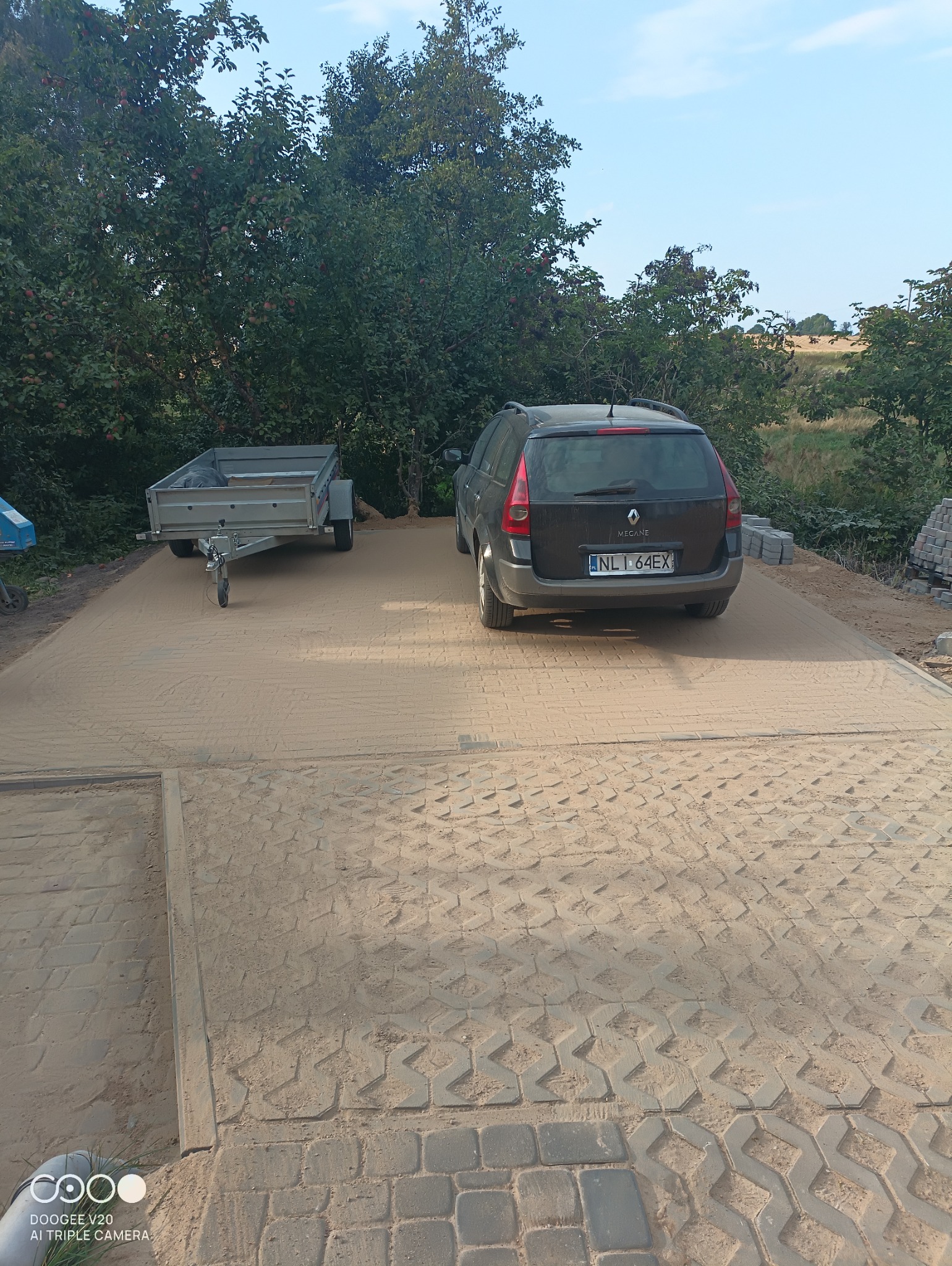 Przygotowany podjazd z podsypką piaskową pod kostkę brukową, zaparkowany samochód i przyczepka w tle.