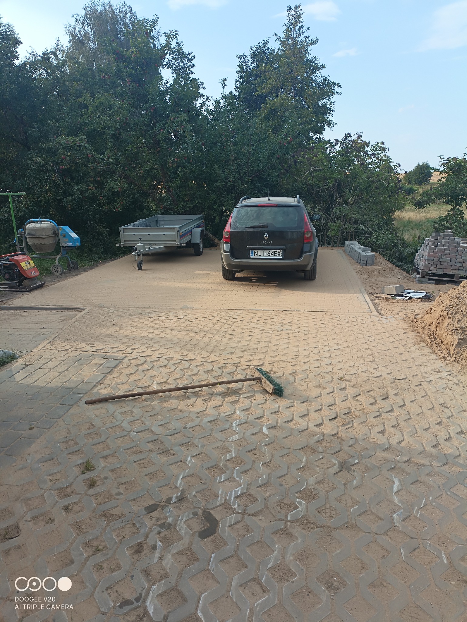 Ułożona ażurowa nawierzchnia z kostki betonowej, zasypana piaskiem, z widocznym samochodem osobowym, przyczepką, betoniarką i stertą kostki w tle, oraz leżącą szczotką.