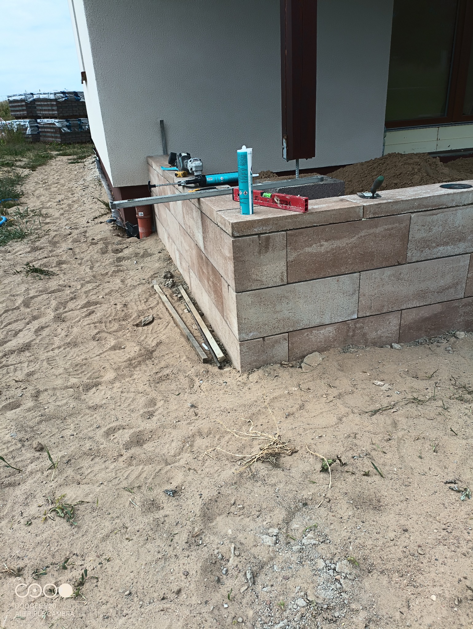 Budowa murku oporowego z bloczków betonowych: widoczne narzędzia, poziomica, klej montażowy, szlifierka kątowa, oraz ułożone bloczki i podsypka z piasku.