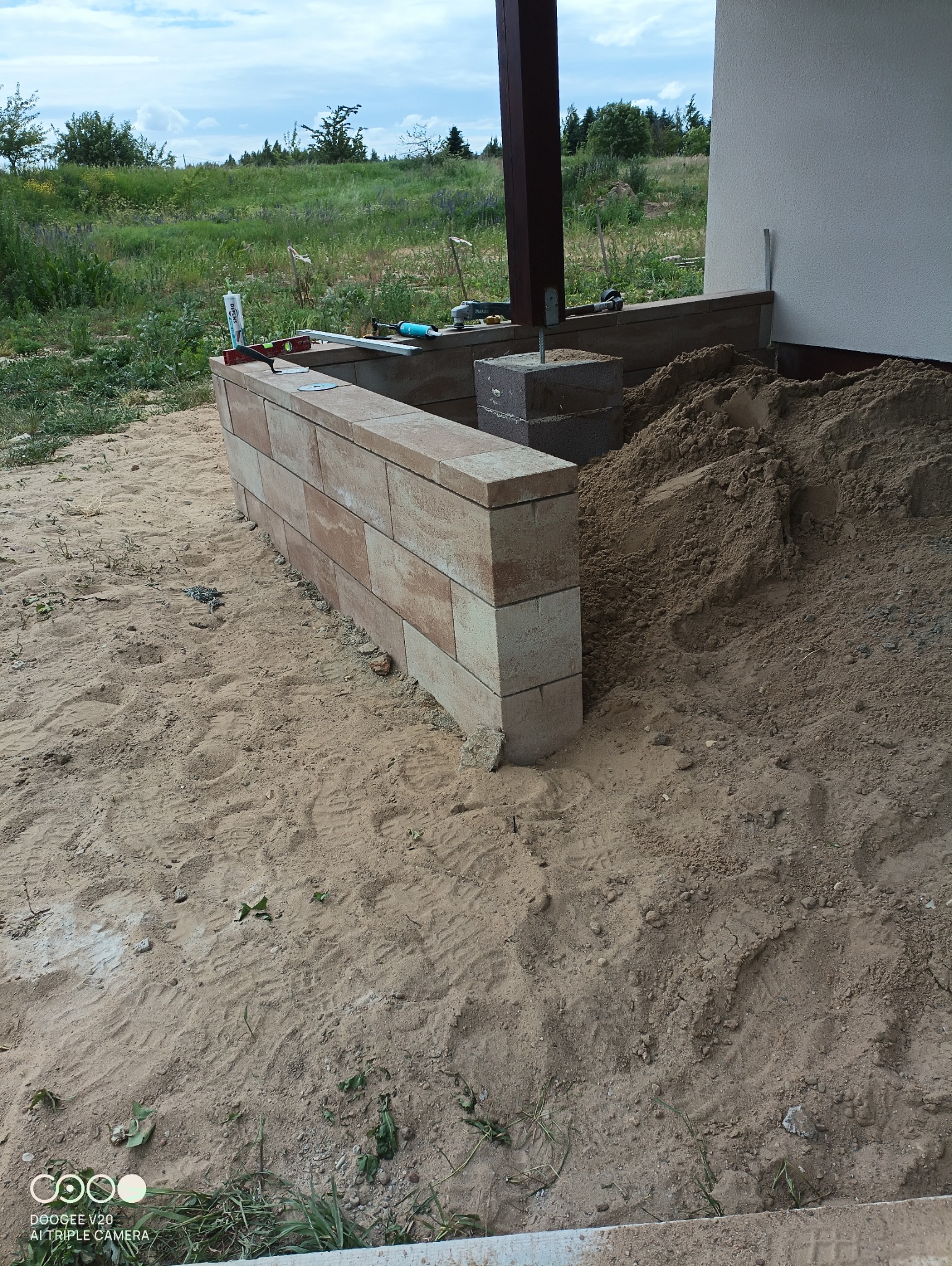 Wznoszenie murku z kostki brukowej przy budynku, widoczny piasek, narzędzia i elementy konstrukcyjne drewnianej wiaty.