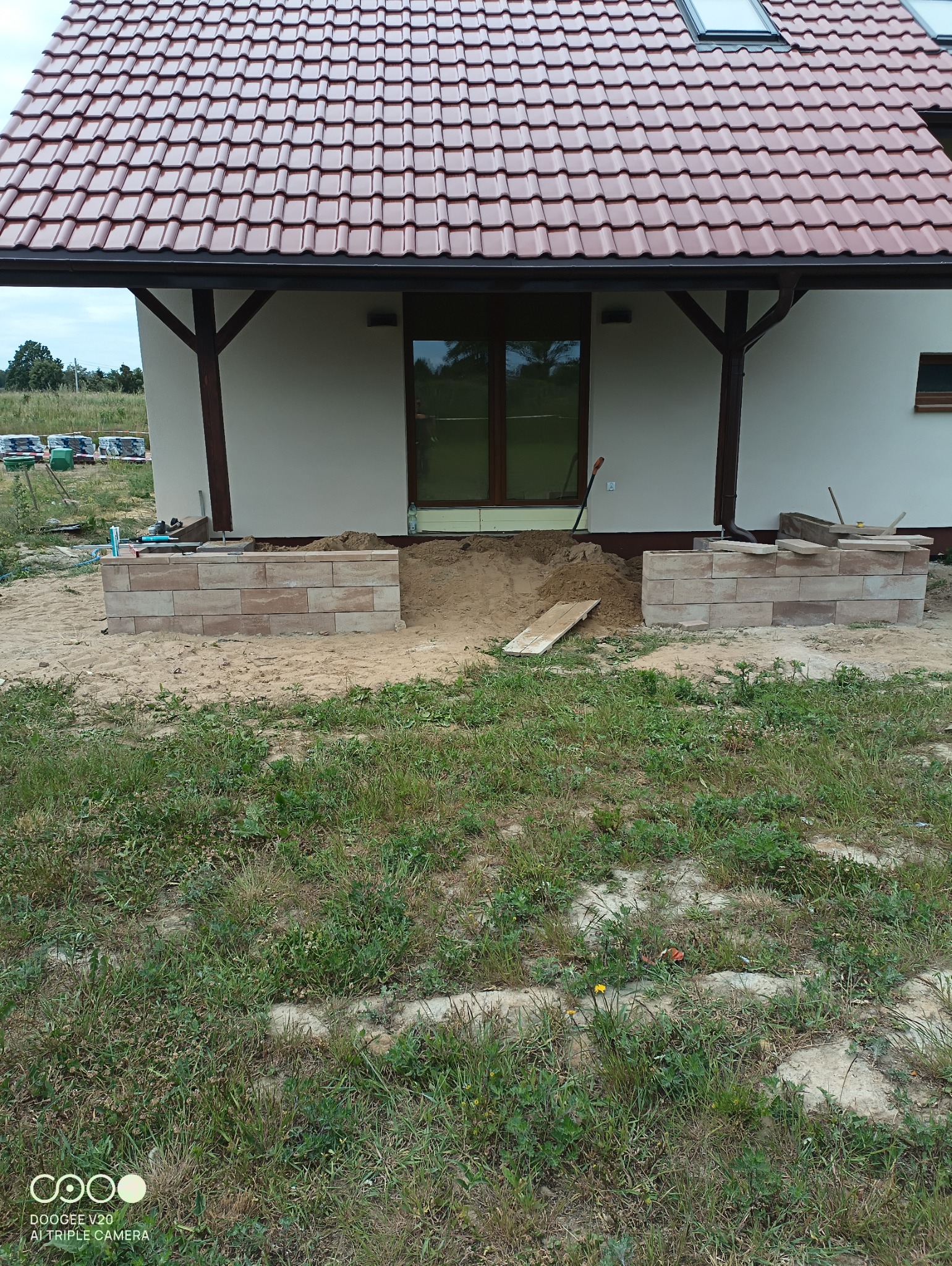 Przygotowanie podłoża i wznoszenie murku z kostki brukowej przed wejściem do domu z brązową stolarką okienną i dachem z czerwonej dachówki, widoczna łopata i zapasy kostki w tle.