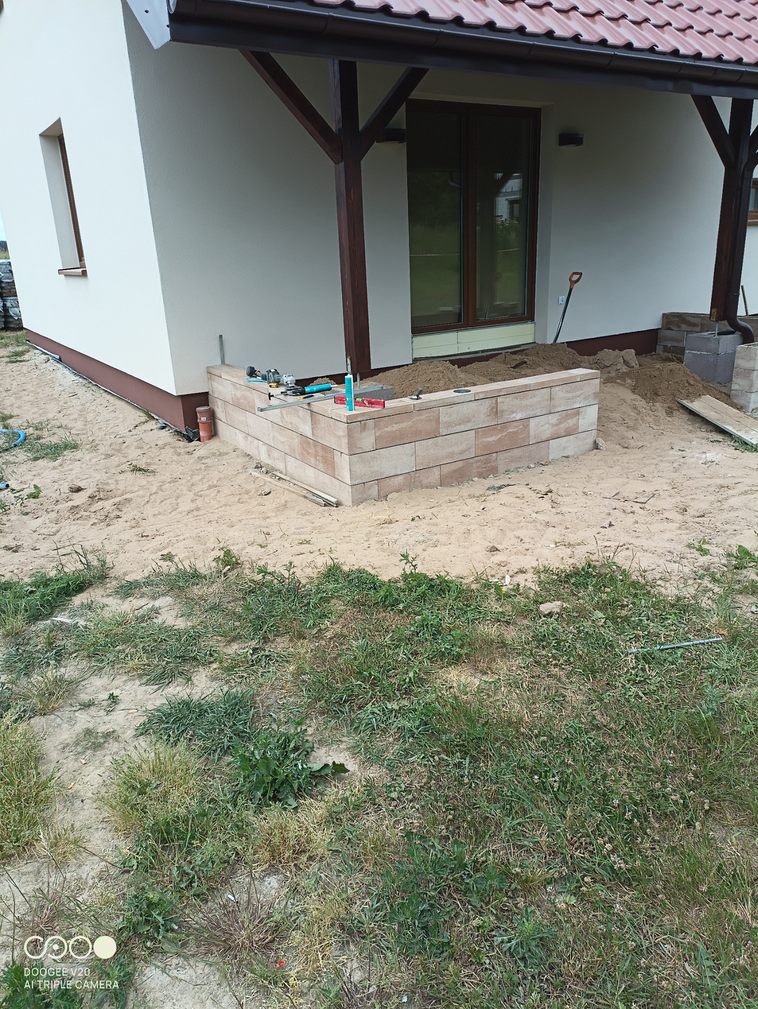 Budowa murku z beżowej kostki przy budynku, widoczne narzędzia i piasek, trawa w tle.