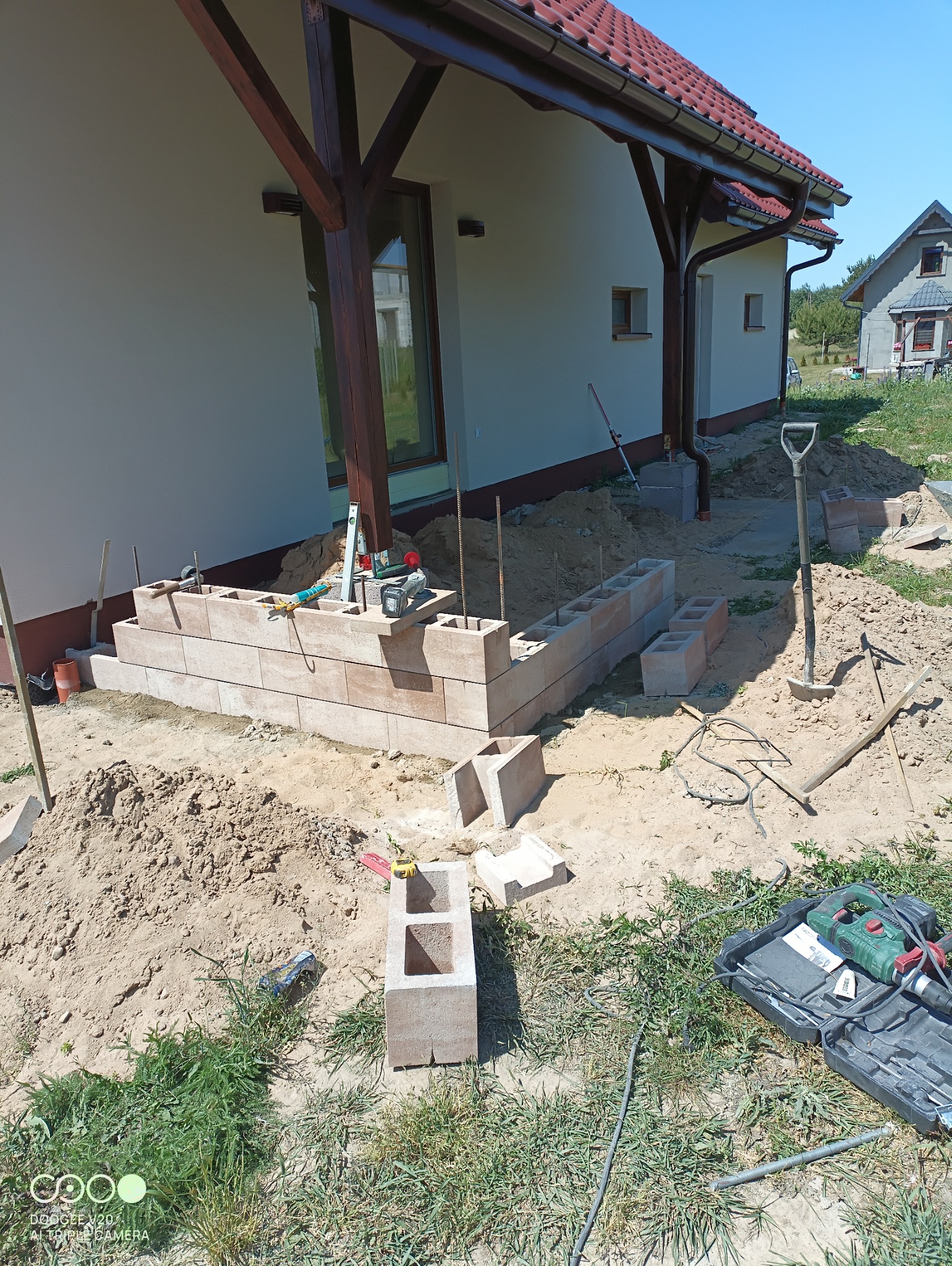 Budowa murku z betonowych bloczków przy domu z drewnianą konstrukcją dachu, widoczne narzędzia budowlane i poziomica.