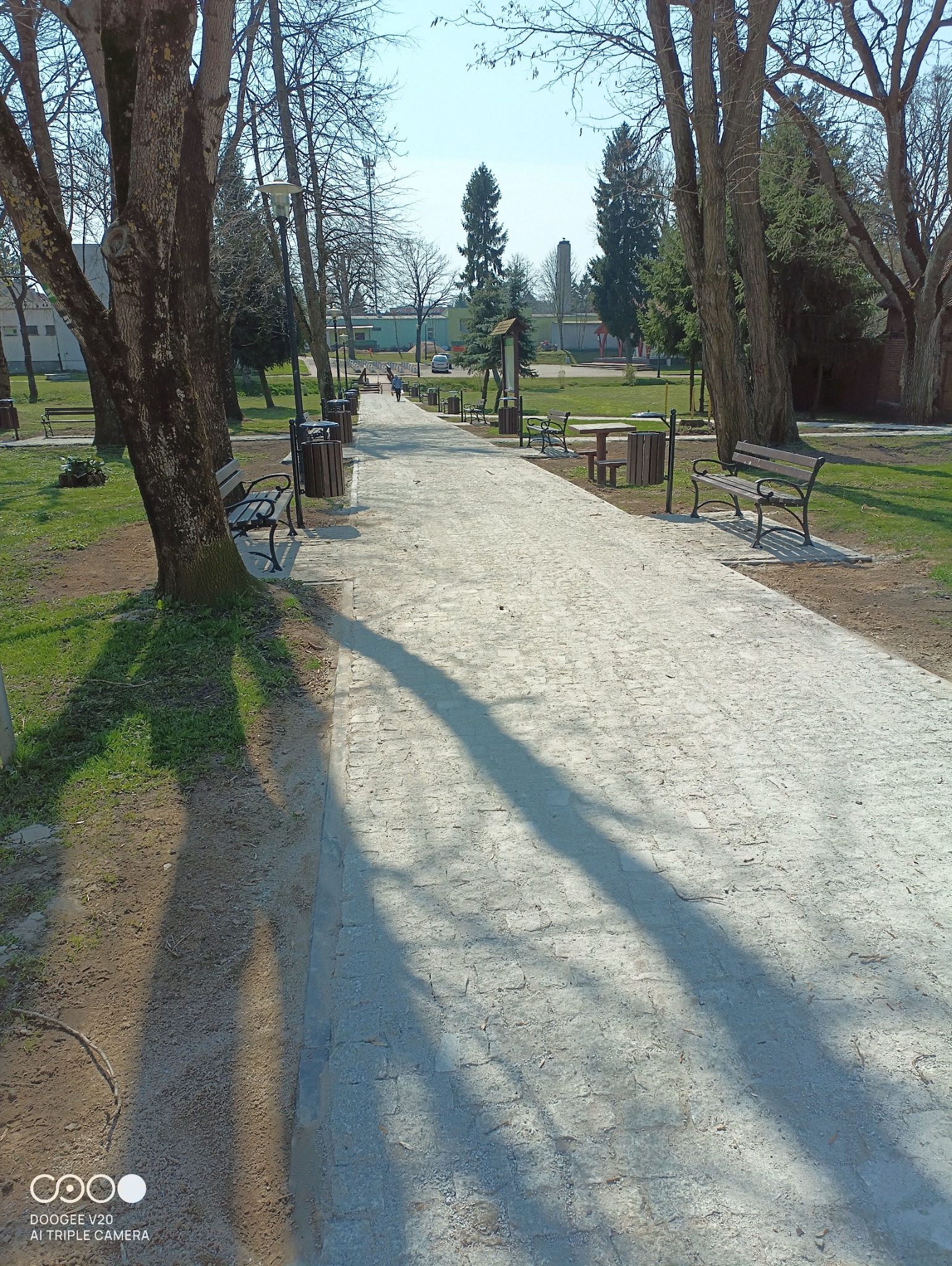 Parkowa ścieżka z jasnej kostki, z rzędami drzew po bokach, ławkami i koszami na śmieci. W tle widoczny budynek i komin.