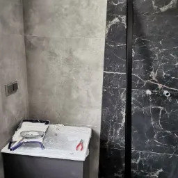 Wnętrze łazienki w trakcie wykańczania: szare płytki na ścianach, czarna kabina prysznicowa z marmurowym wzorem, narzędzia glazurnicze i wiadro z zaprawą na umywalce przy ścianie z włącznikiem...