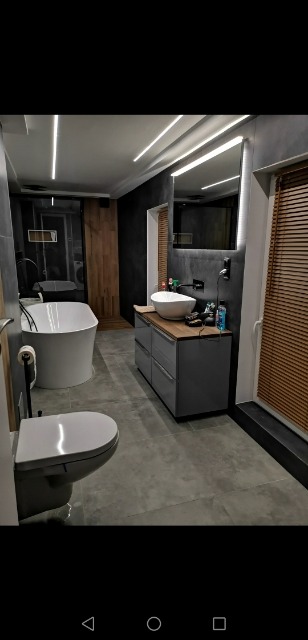 Nowoczesna łazienka z szarą armaturą, wolnostojącą wanną i podświetlanym lustrem. Widoczne drewniane akcenty i rolety w oknie.