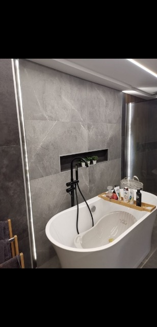 Nowoczesna łazienka z wanną wolnostojącą, szarymi płytkami imitującymi marmur, podświetleniem LED i wnęką dekoracyjną z roślinami.