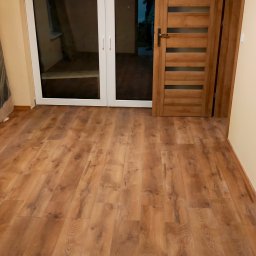 Ihrhause - Pokój po remoncie z drewnianą podłogą, białymi drzwiami balkonowymi i drewnianymi drzwiami wewnętrznymi z przeszkleniami, widok na częściowo wyposażone wnętrze.